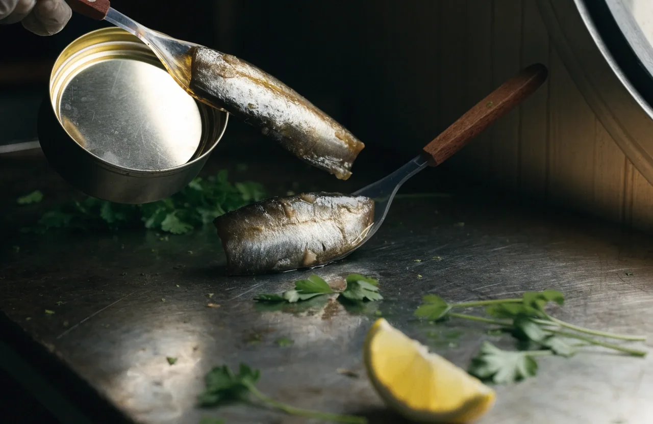 An oily fish fillet on baking parchment with a lemon half, side-lit against a dark tray