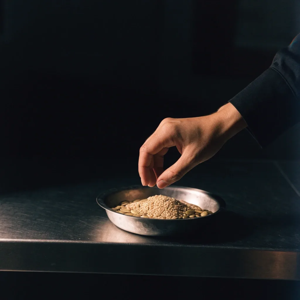 A hand reaching into a steel dish of mixed seeds and nuts at the edge of a dark kitchen table at night